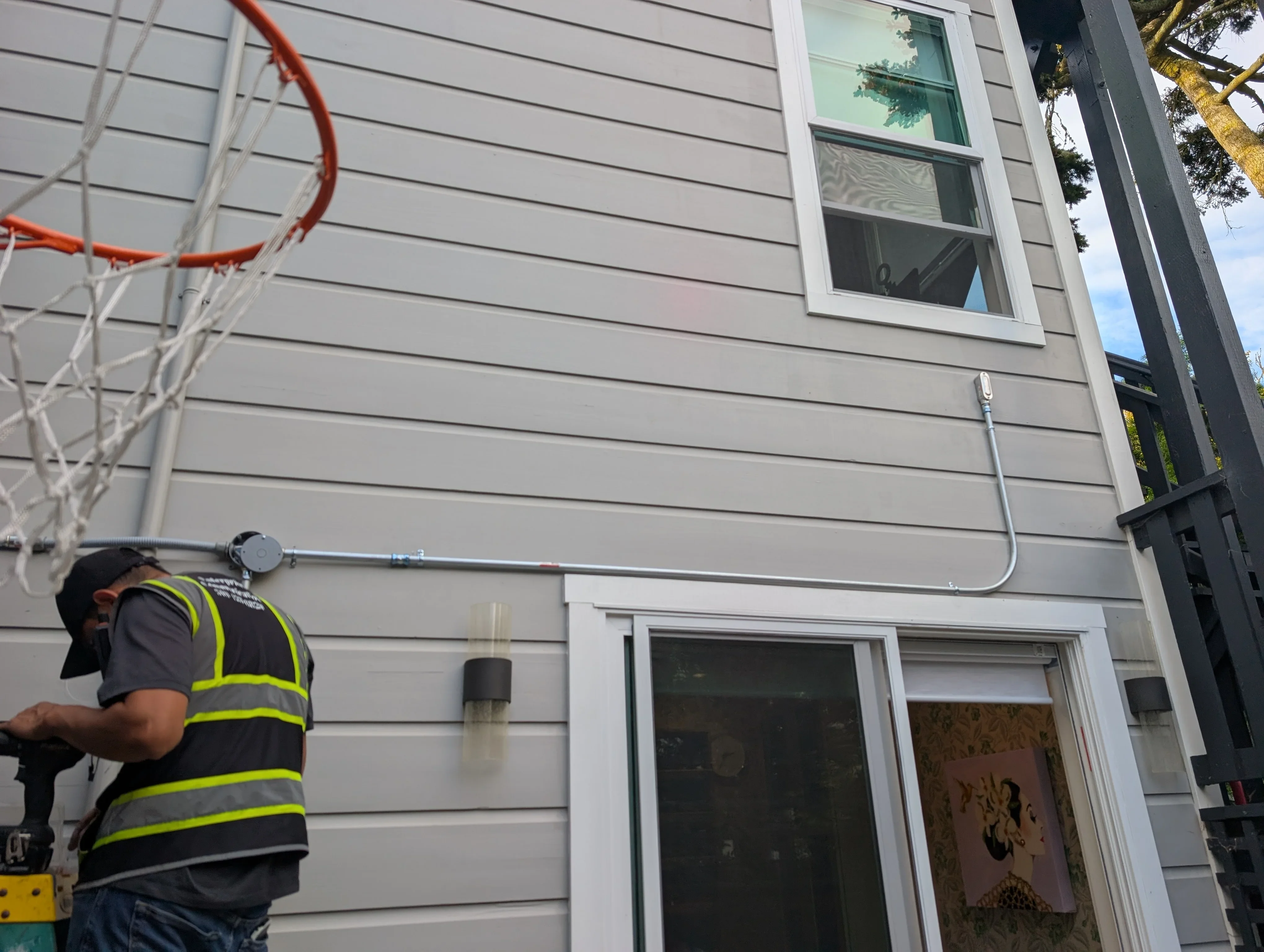 Technician installing gray telecommunications conduit on residential siding with white-trimmed windows